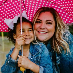 foster carer and fostered child smiling wide at the camera while under an umbrella
